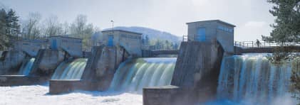 Blick auf ein Wasserkraftwerk. Vier Wasserströme gelangen zwischen vier Mauern mit einem Gebäude darauf durch. Eine Brücke führt über die Wasserfälle. darüber.