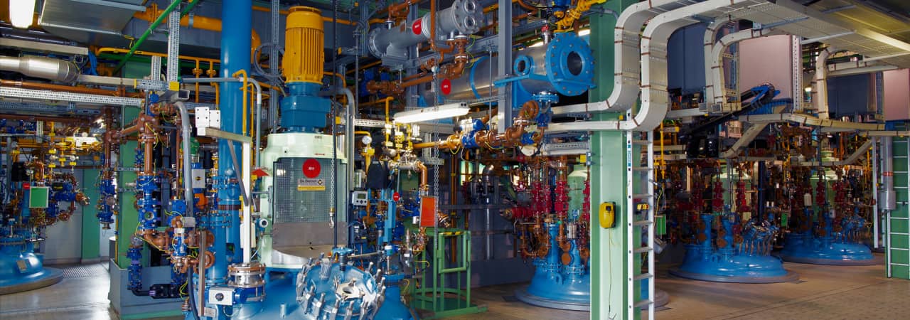 Halle mit vielen Metallrohren in blau, Grün, gelb und orange und blaue Maschinen. Das Bild wirkt sehr komplex.