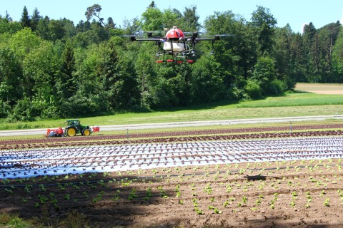 Eine rote Drohne schwebt über einem Feld, auf welchem Pflanzen wachsen. Im Hintergrund sieht man einen Traktor, der das Feld bestellt.