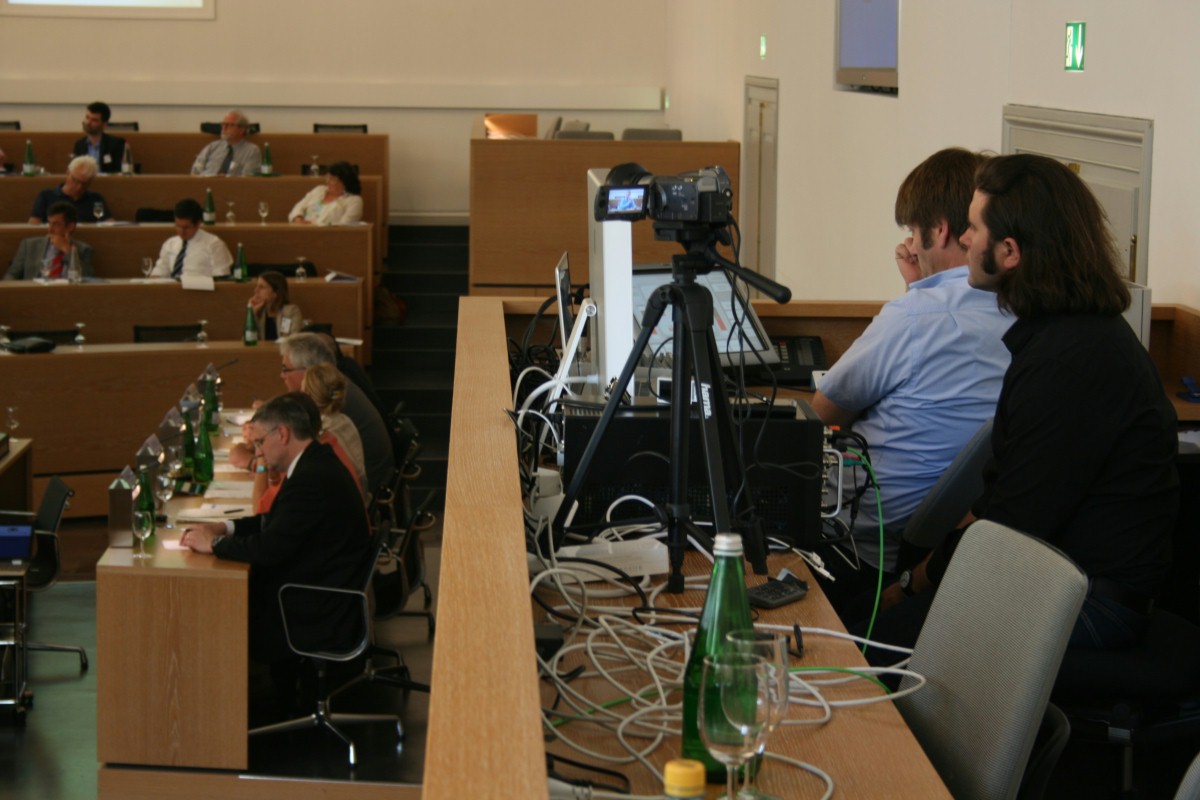Bild von der Medienplätzen aus aufgenommen: Rechts sitzen zwei Techniker vor Kamera und Computer und links im Hintergrund findet eine Debatte statt