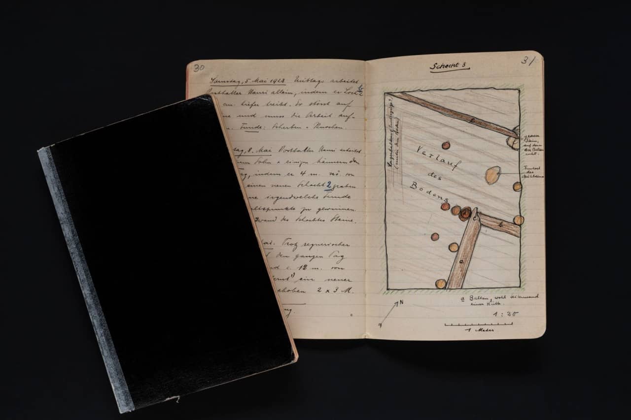 Ein aufgeschlagenes Tagebuch rechts mit der Zeichnung einer archäologischen Grabung, rechts daneben ein schwarzes Notizbuch.
