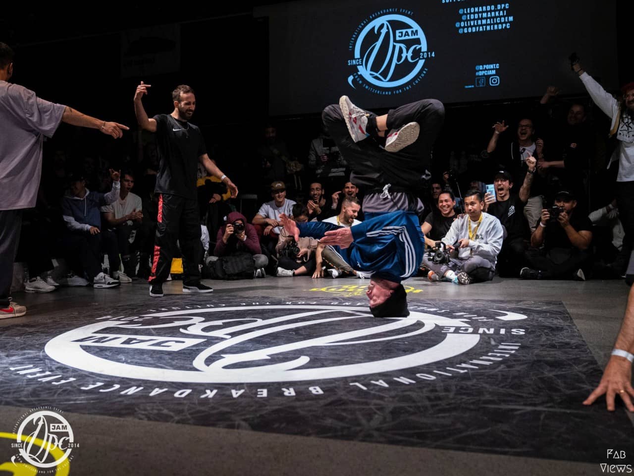 Ein Mann breakt an einem Breakdance-Event, Männer neben ihm und das Publikum feuern ihn an. 