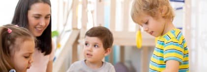 Eine Kindergartenlehrerin kniet neben drei Kindergartenkinder, die an einem Tisch spielen und basteln.