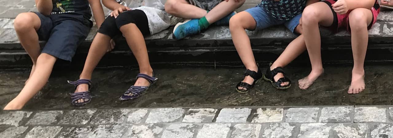 Kinder und Erwachsene sitzen an einem kleinen Stadtkanal, baden Ihre Füsse und essen dazu Eis.